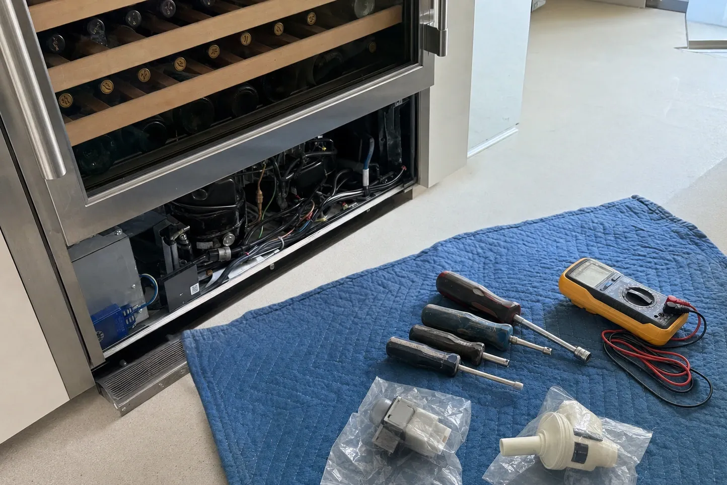 Wine cooler with the glass door open on racks of bottles, plinth panel off and the compressor exposed, tools on a blanket