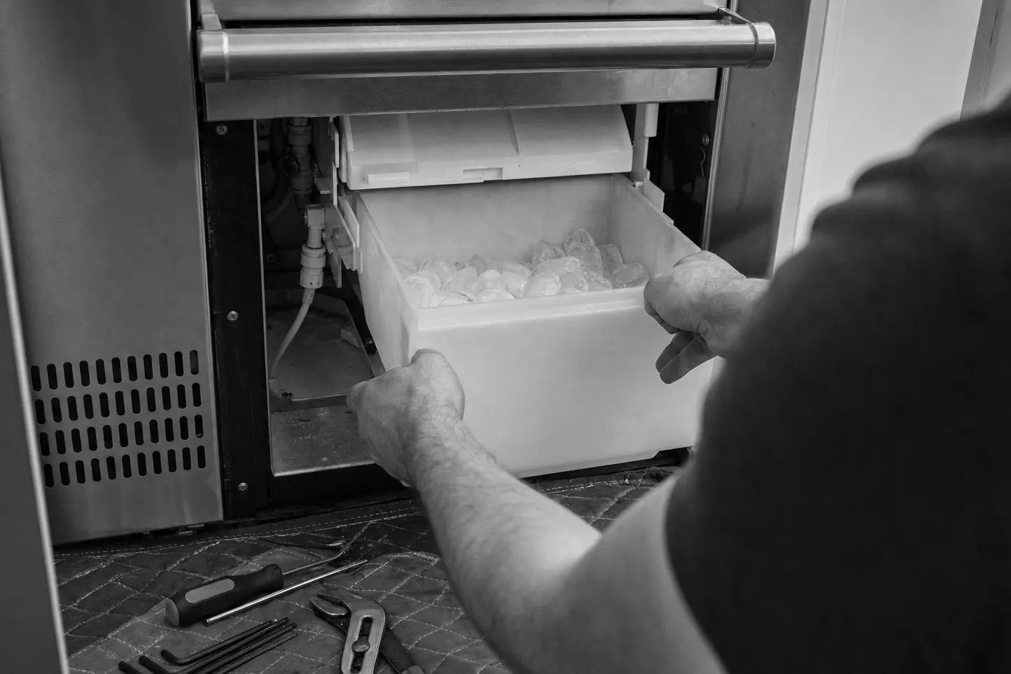 Black and white view of the ice storage bin, full of cubes, being drawn out of an undercounter ice maker with the water line behind it