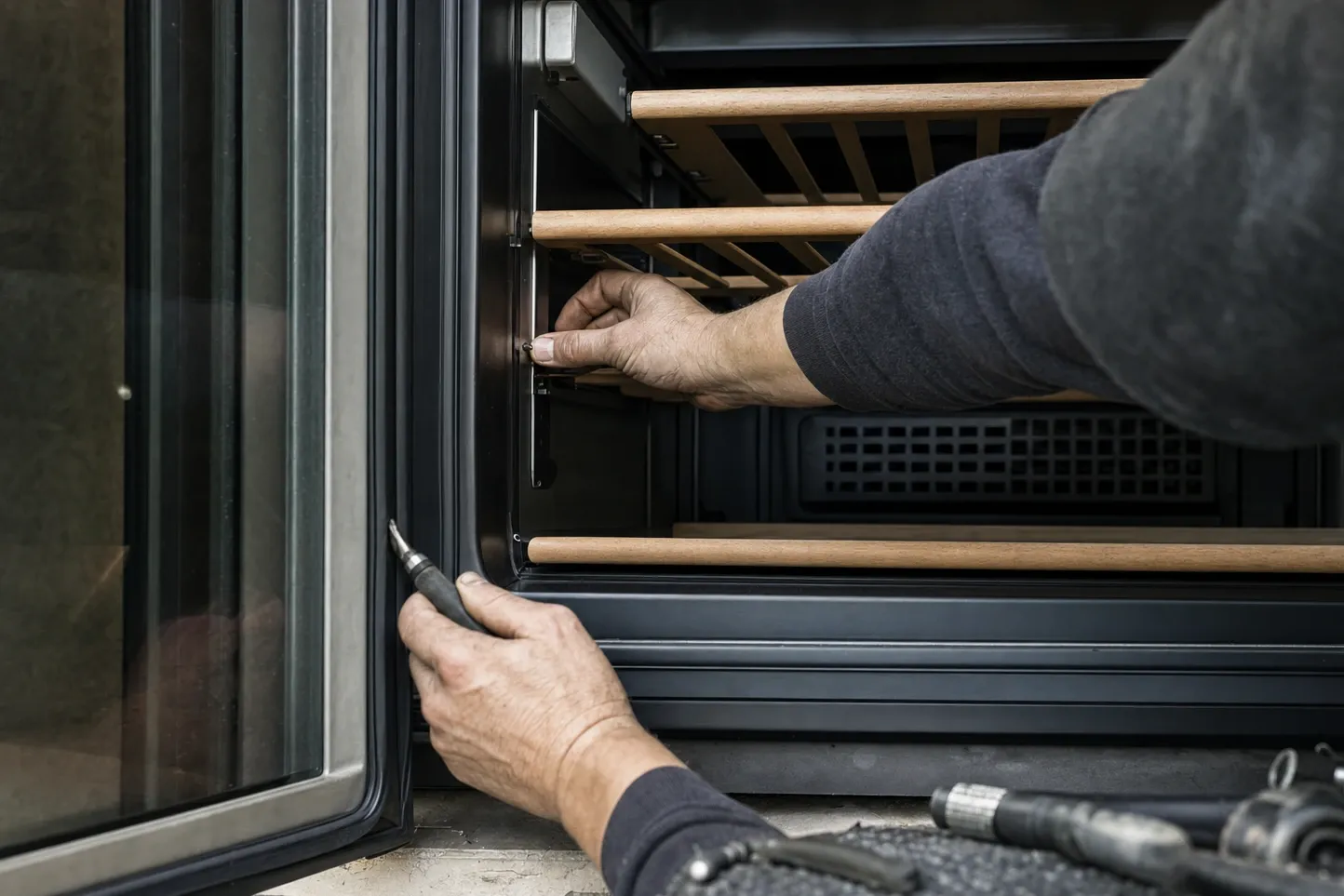 Hands at the open door of a dark undercounter unit with bare wooden shelves, the rear vent grille visible behind them and a small driver at the door frame